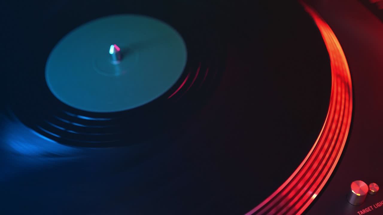 The player vinyl turntable close-up on a background of red and blue lights. The young man's hand smoothly places the needle on the rotating plate. DJ at a youth student's party
