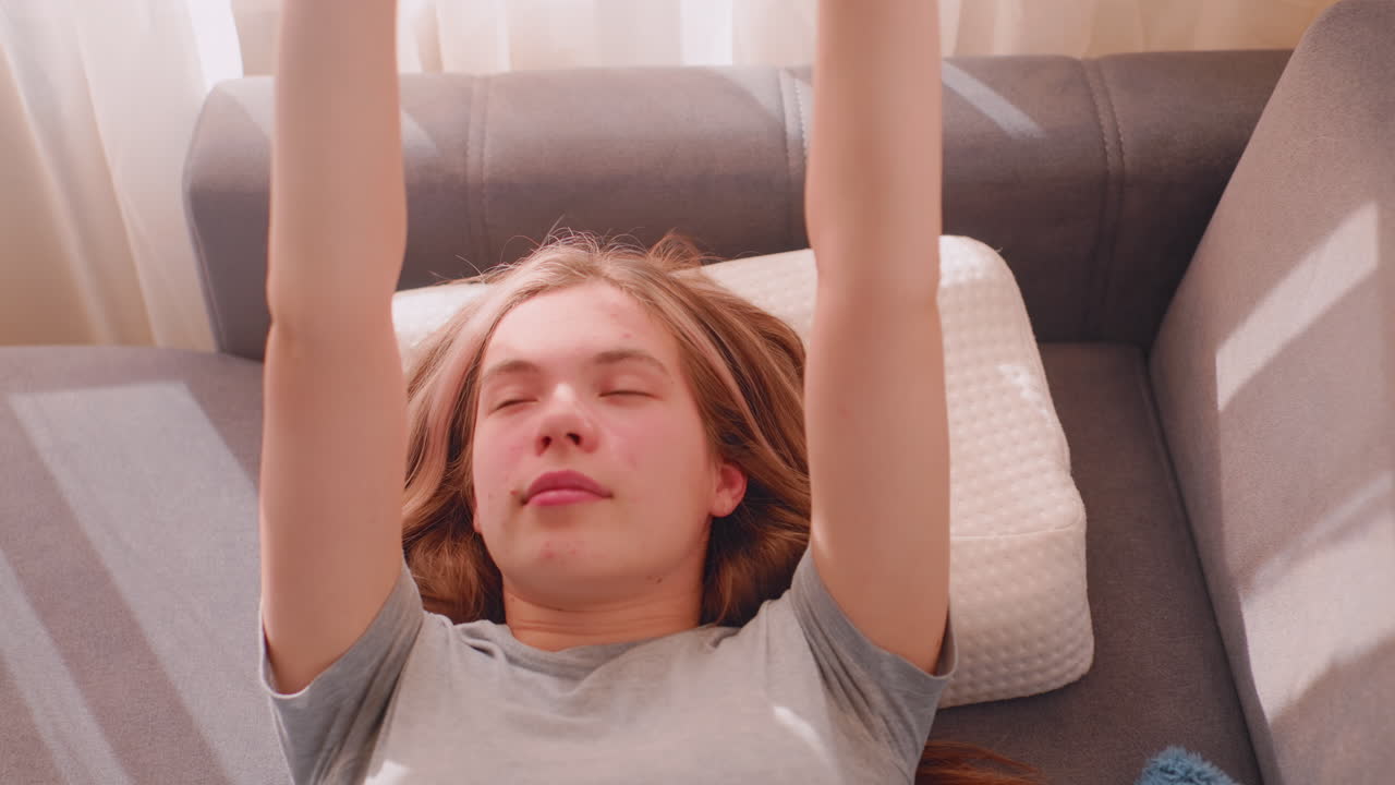 Top down view of student lying on couch with blanket, slowly waking up and stretching shoulders in soft daylight, relaxed expression showing peaceful morning moment after sleep