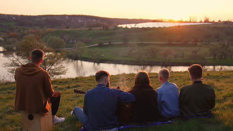 group of friends sitting on the slope of hill and playing musical instruments. young company enjoying sunset and summer evening.