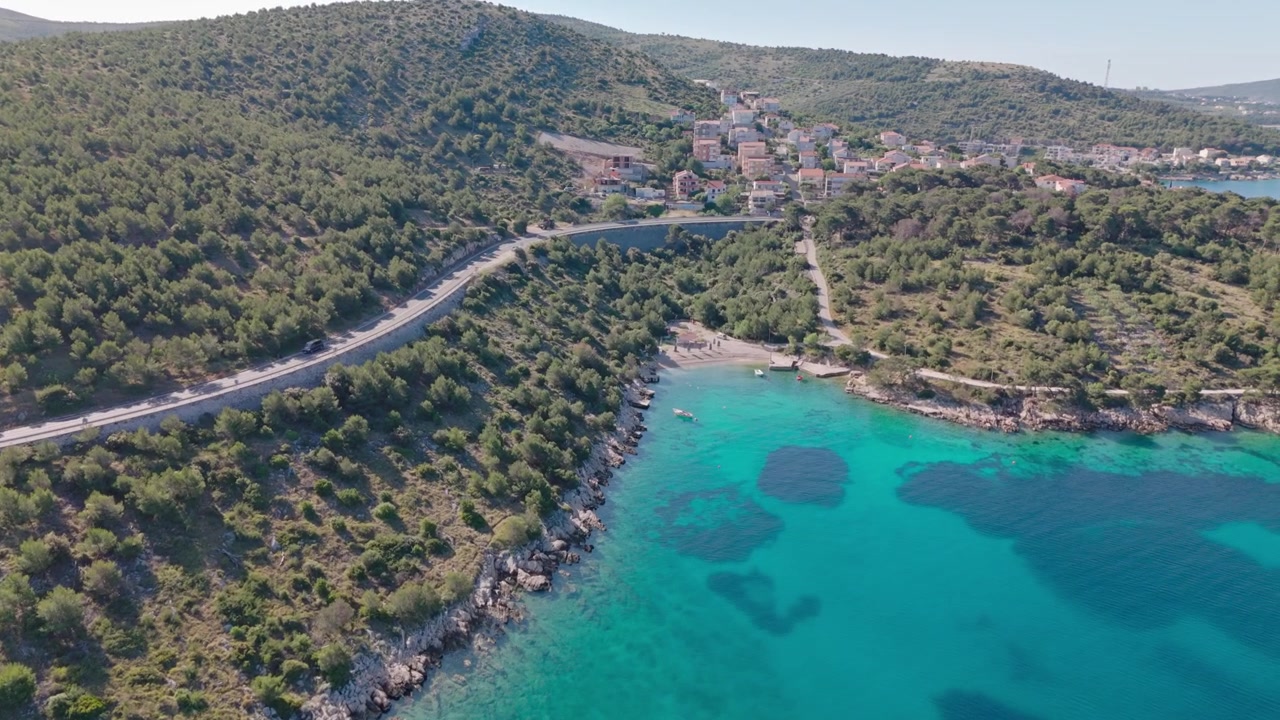 Drone che vira con una curva morbida lungo la Magistrala nella Contea di Sebenico e Tenin, rivelando una luminosa cala turchese e la frastagliata costa adriatica sotto la luce estiva.
