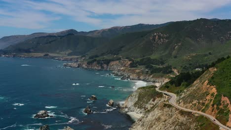 wide aerial view of a winding curvy highway road along route 1 on the coast of big sur california with large mountain cliffs and waves crashing into the rocks below