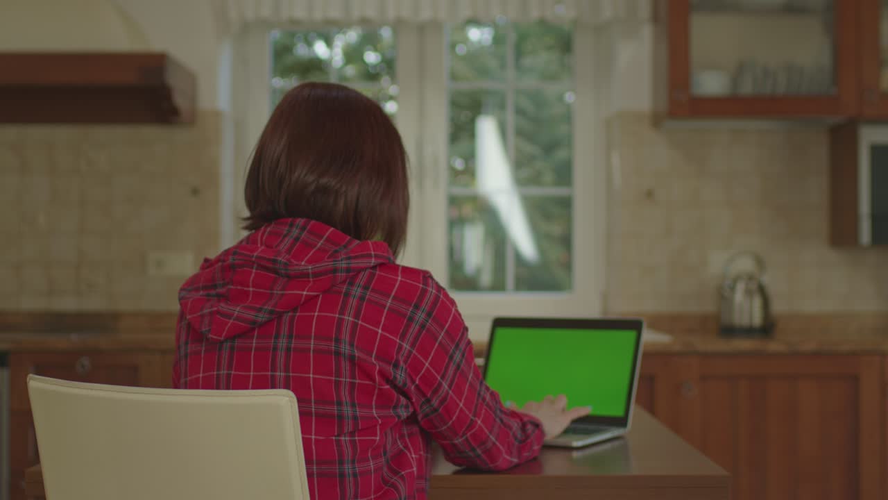 Back view of woman working on laptop with green screen sitting in the kitchen.