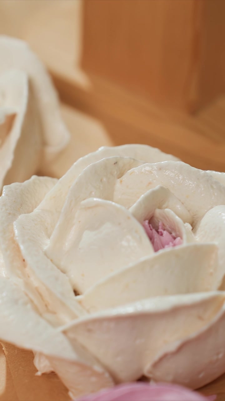 Primo piano di gelato bianco a forma di rosa con un dettaglio centrale rosa. La consistenza cremosa e i dettagli dei petali sono evidenziati. Lo sfondo include elementi sfocati che suggeriscono una cucina interna.