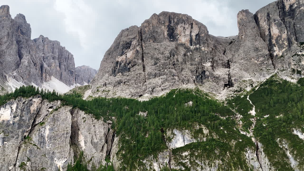 Vista aerea con drone di un'enorme scogliera verticale di calcare che si erge sopra una fitta foresta alpina. L'imponente parete rocciosa domina il paesaggio montuoso delle Dolomiti.