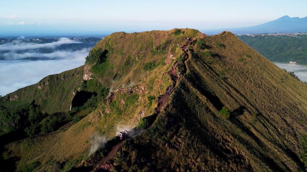 Escursionisti che scalano il vulcano Monte Batur a Bali lungo un ripido sentiero di vetta sopra le nuvole, con fumo vulcanico che si diffonde attraverso un paesaggio aspro e ad alta quota. Ripresa con drone in avvicinamento.