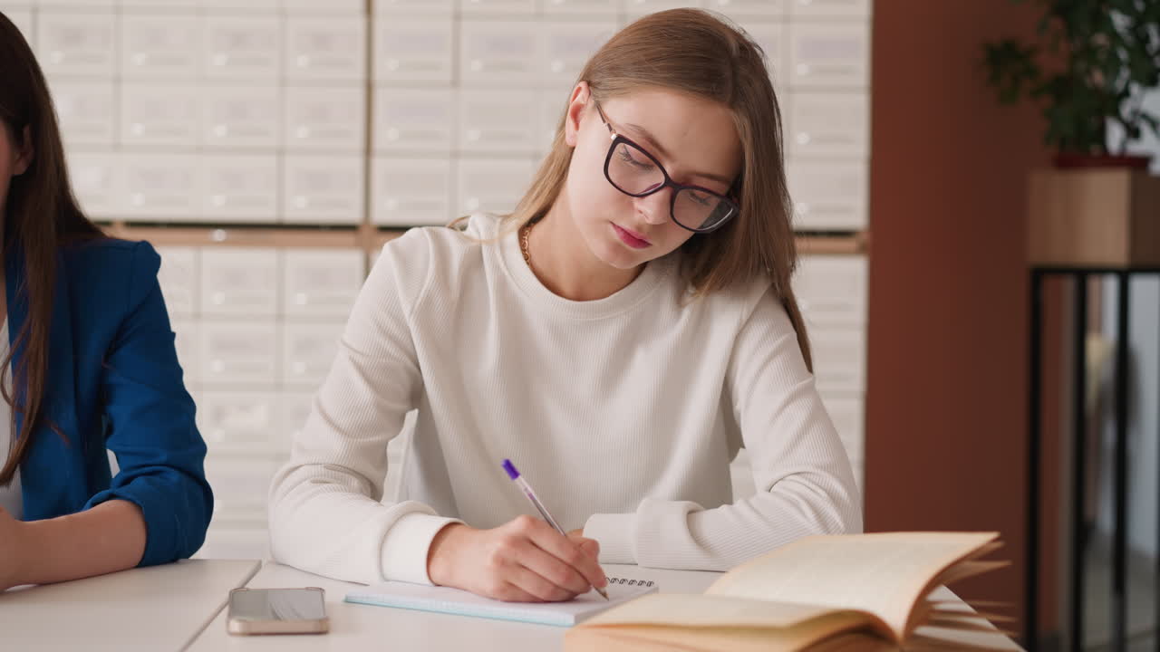 una joven rubia escribe notas en la biblioteca de la universidad. una estudiante con gafas se sienta en la mesa preparándose para la clase usando un libro educativo. atención durante el estudio