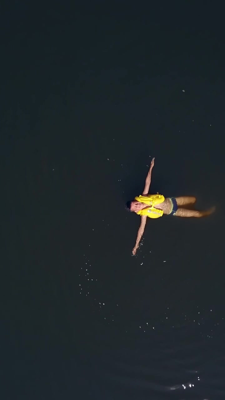 Happy little child in yellow life vest swimming alone on his back in the calm river. Teenage boy wearing safety waistcoat cheerfully swimming. Aerial view. Motion bottom up Vertical video