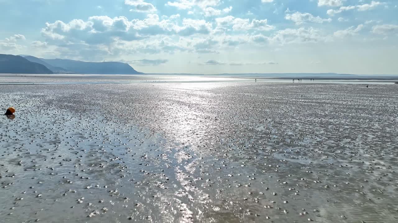 한 무리의 관광객들이 갯벌 지역의 말라붙은 진흙 바닥을 가로질러 걷고 있다. 햇빛이 진흙과 물 위로 반짝이며 웨일즈 산맥을 배경으로 하고 있다.