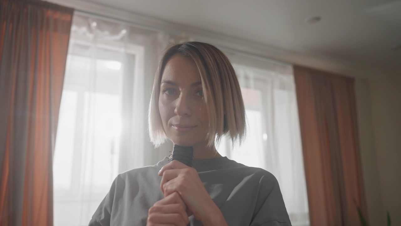 Mujer blanca cantando en un micrófono con luz de ventana, contraluz suave y cortinas cálidas, corte bob, top corto casual, cepillo juguetón usado como micrófono, ambiente íntimo de estudio en dormitorio, estado de ánimo tierno y romántico