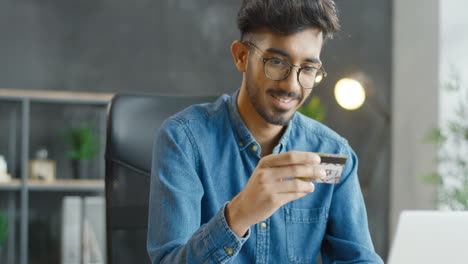 close up view of arabic young man in eyeglasses sitting at desk, holding credit card and shopping online on laptop computer