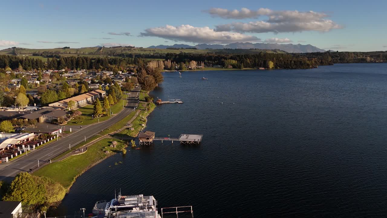 Scenic 4K aerial drone footage flying backwards along the Te Anau waterfront. Views of the town, pier, deep blue lake, and distant mountains in Fiordland, New Zealand