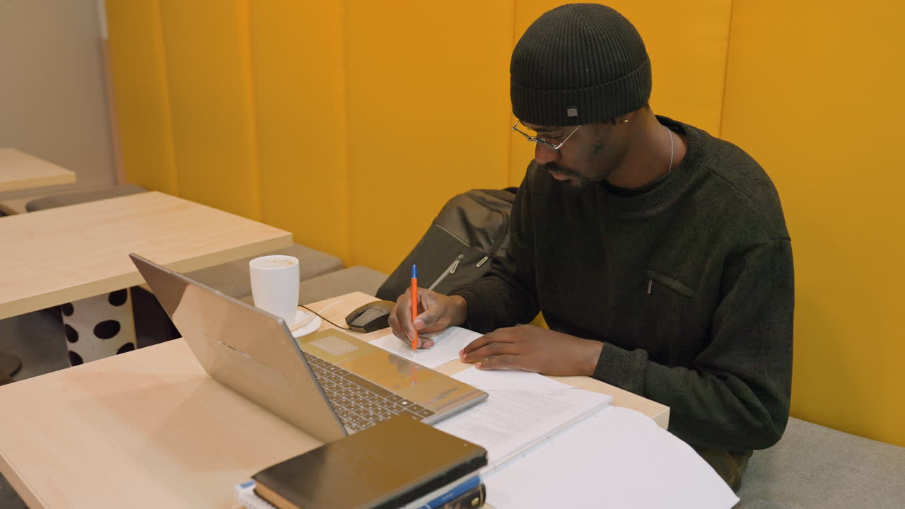 Studente di colore che studia alla scrivania con il laptop, che prende appunti con una penna, pila di libri di testo e quaderno aperto accanto al laptop, pannelli acustici gialli, tazza in ceramica, atmosfera di studio tranquilla in coworking, produttività serale