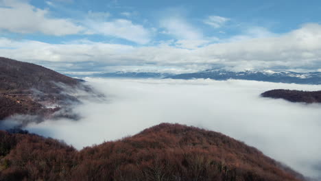 A view of land with trees and snow covered mountains in the distance There is also a layer of clouds below The sky has clouds and patches of blue