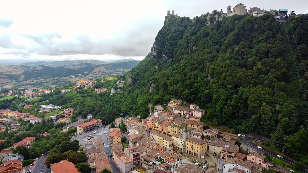 Aerial view pulling back over San Marino castle, old town buildings, forested hills and cable car, scenic mountain backdrop with historic charm