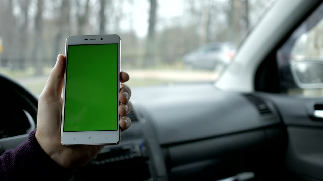 una mano sosteniendo un teléfono inteligente con pantalla verde en blanco en el coche para la dirección, masaje, ubicación, negocio. hombre se sienta en un coche moderno y trabaja en teléfono inteligente - pantalla verde - primer plano. llave de croma.