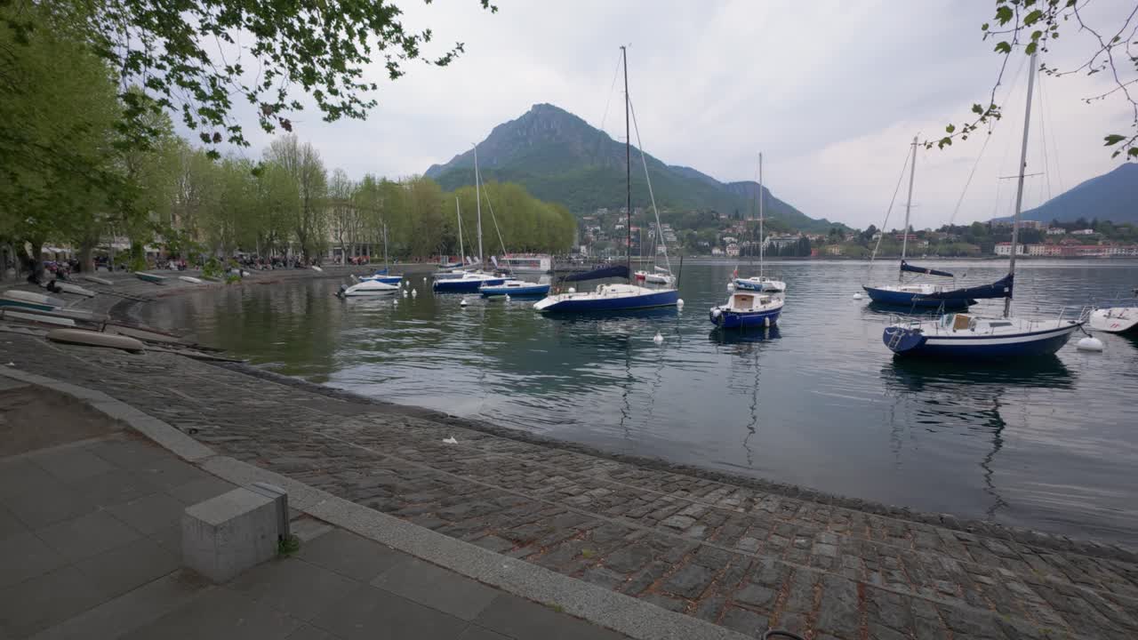 Ampia inquadratura panoramica di barche a vela ancorate sul Lago di Como a Lecco, Italia, circondate da alberi rigogliosi, acqua tranquilla e montagne sotto un cielo leggermente nuvoloso. Nessuna persona visibile, luce naturale.