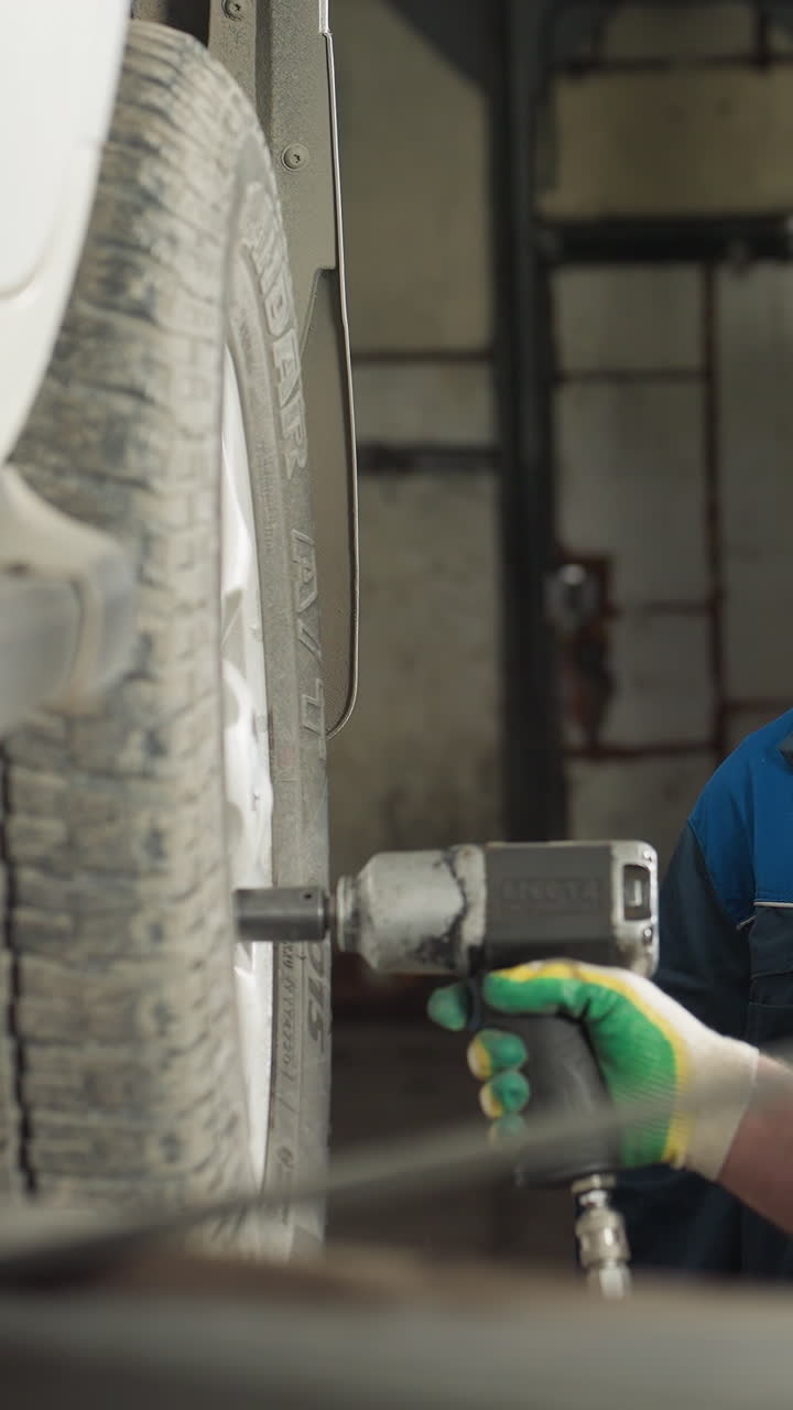 dos ingenieros automotores que trabajan en un taller mecánico, uno usando una herramienta neumática para quitar los pernos de las ruedas de un coche suspendido por un ascensor, mientras que el otro observa con una sonrisa, tocando su cabeza