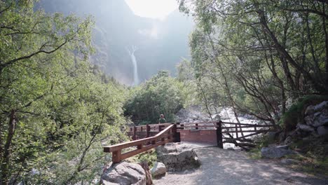 Mujer-Haciendo-Turismo-Con-Un-Perro-Cruzando-Un-Puente-De-Madera-Con-Cascada-Mardalsfossen-Al-Fondo-En-Noruega