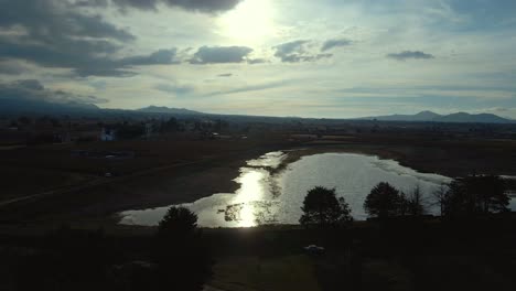 Panoramic-aerial-orbit-sun-reflecting-on-a-lake,-lone-car-in-the-natural-landscape-of-Almoloya-in-Hidalgo-Mexico