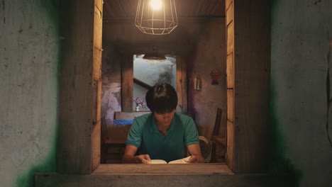 mirando a través de una ventana abierta a un joven hombre asiático lee un libro en un escritorio en una habitación rústica