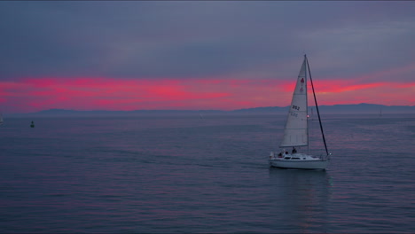 sailboat on ocean with pink and orange sunset looking toward the skyline with island mountains