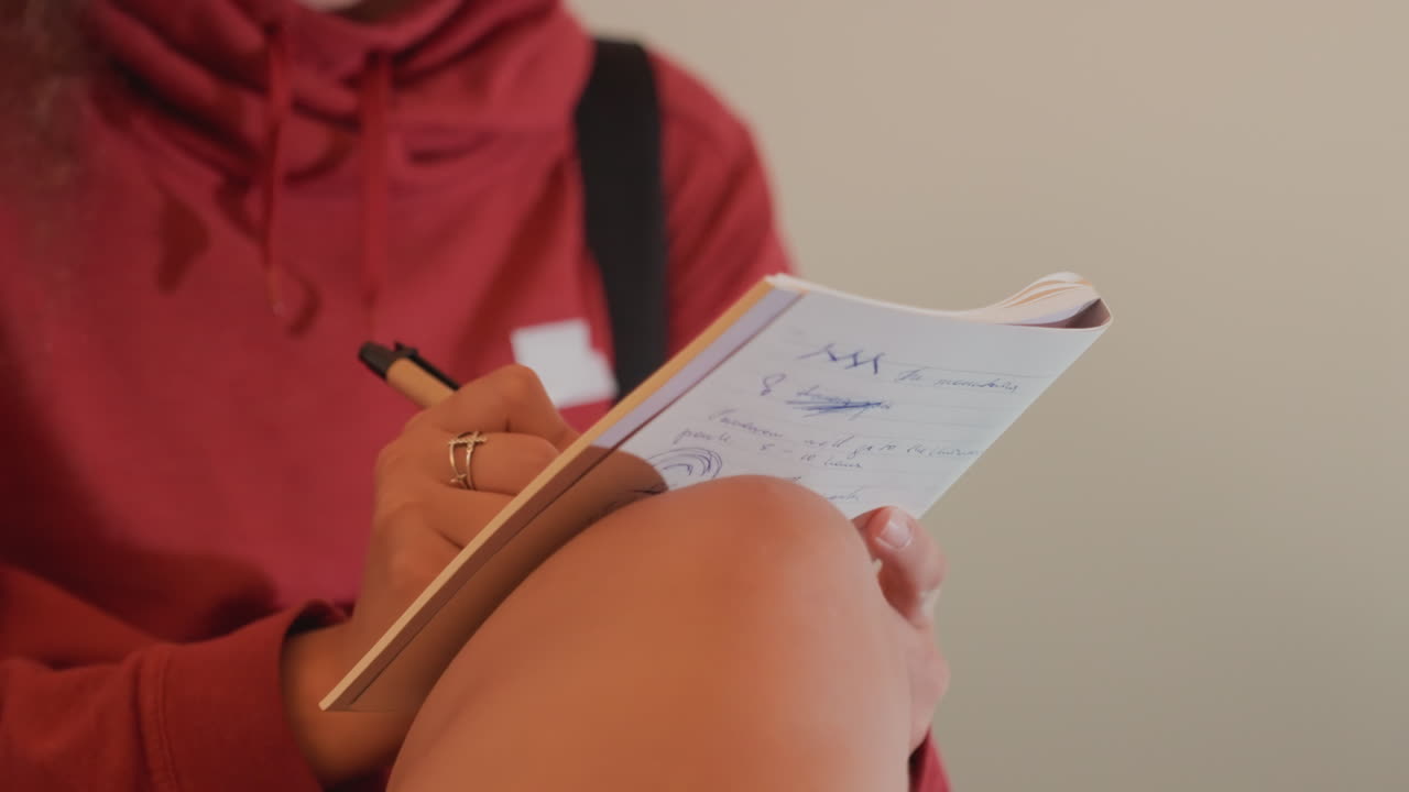 Young woman writing in notebook on lap, red hoodie and rings, pen moving across lined pages, warm indoor bokeh lights, quiet reflective mood, personal notes and sketches captured in soft film grain
