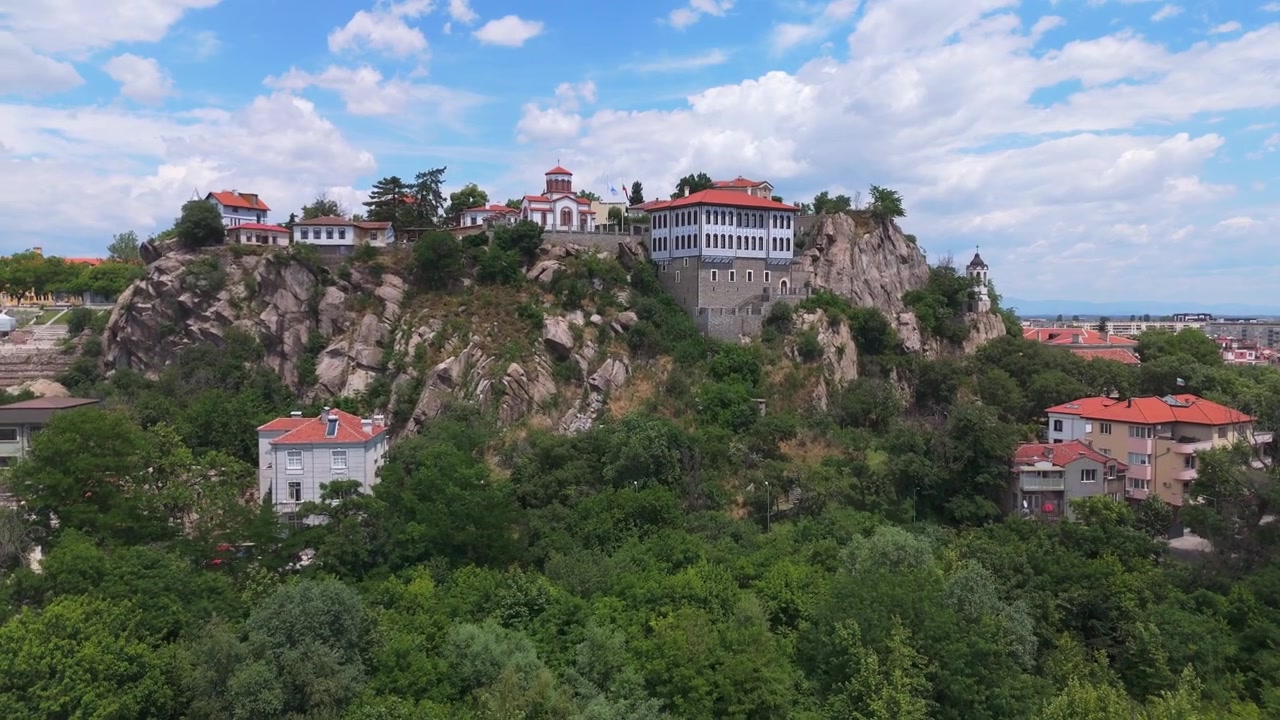 Tomada aérea ampla de drone aproximando-se das ruínas da fortaleza na colina verde de Nebet Tepe, exibindo marcos arquitetônicos históricos no bairro da cidade velha de Plovdiv, Bulgária