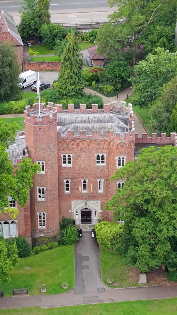 Cinematic close vertical aerial of Hertford Castle’s front as the drone reverses and ascends, gradually showing the surrounding greenery, road, and wider Hertfordshire landscape