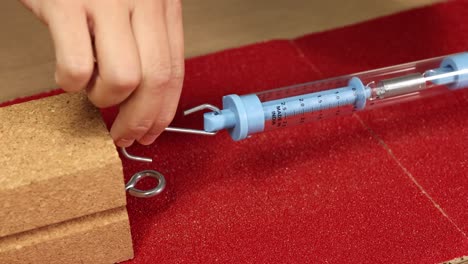 A hand uses a spring scale to measure friction on a red surface with a wooden block