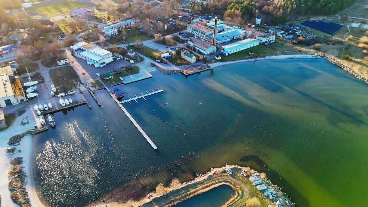 A tranquil coastal harbor features docks, boats, and industrial buildings along a gently curved shoreline, bathed in warm golden light during early evening.
