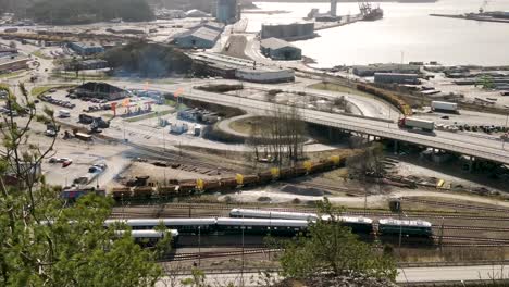 Clip-Aéreo-Del-Tren-De-Carga-Que-Se-Mueve-Lentamente-A-Lo-Largo-De-Las-Vías-En-Medio-De-Una-Carretera-Muy-Transitada-En-Uddevalla-Suecia