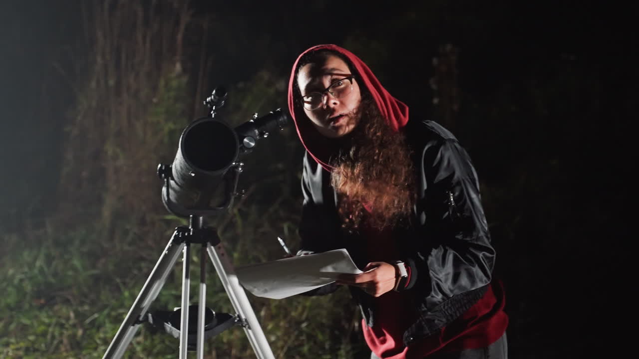 persona con telescopio di notte in un campo, osservatore incappucciato in rosso che calibra il telescopio usando un tablet e carte celesti, espressione concentrata, silhouette barbuta, luce ambientale soffusa, atmosfera da osservatorio rurale in giardino, scientifico
