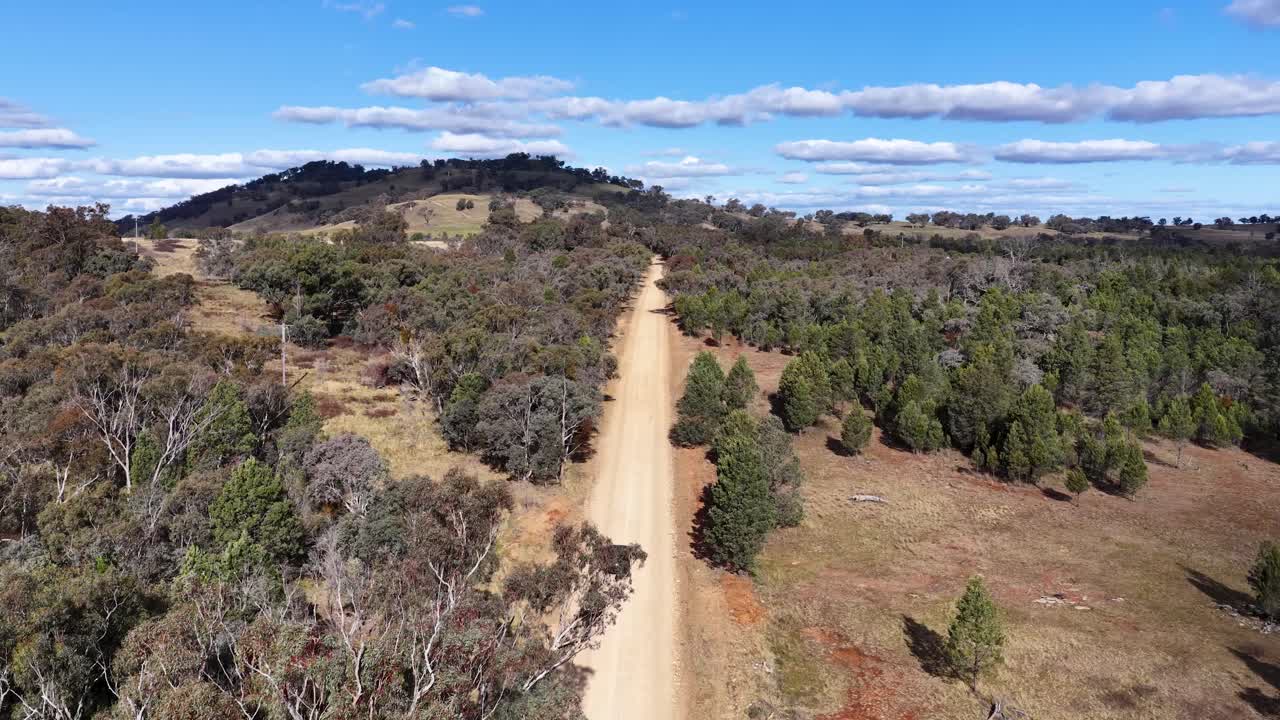 A single vehicle travels along a straight dirt road through a dry, wooded landscape under bright daylight, captured by a steadily moving aerial camera