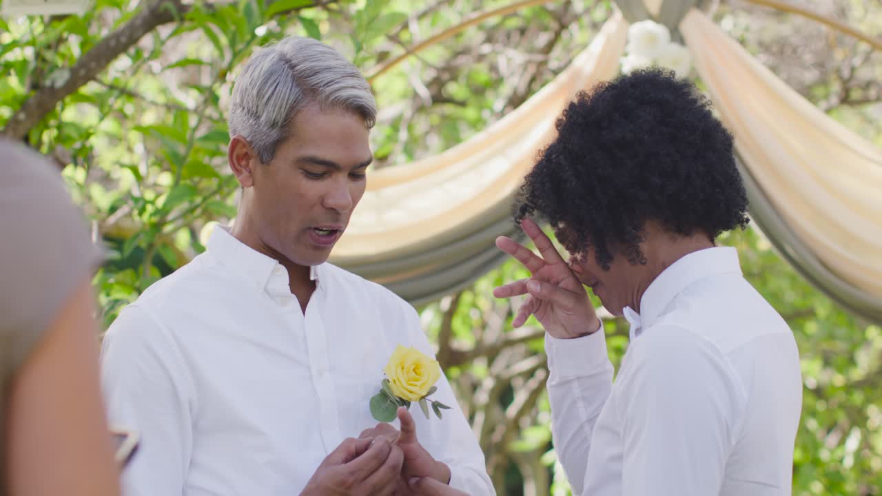 Vertical video: Male pair trading rings at wedding arch, gray man reaching, him tearing up, smiling