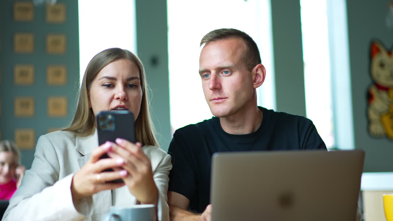 Mid-aged young people sit indoors with laptop in front of them. Lady puts a cup, takes phone and shows something to a man.