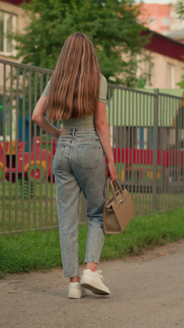 Back view of a woman walking along a path beside an iron fence, holding a beige handbag, her long blonde hair flows down her back as she walks with playground structures visible behind the fence