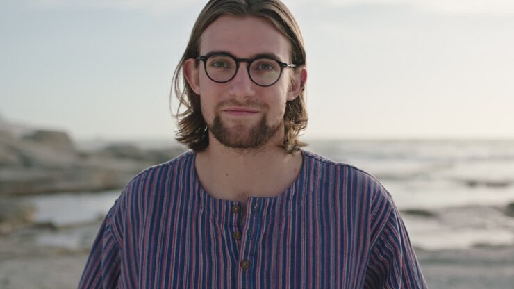 ritratto di un simpatico uomo geek che indossa occhiali sorridente sulla spiaggia che indossa una camicia a righe