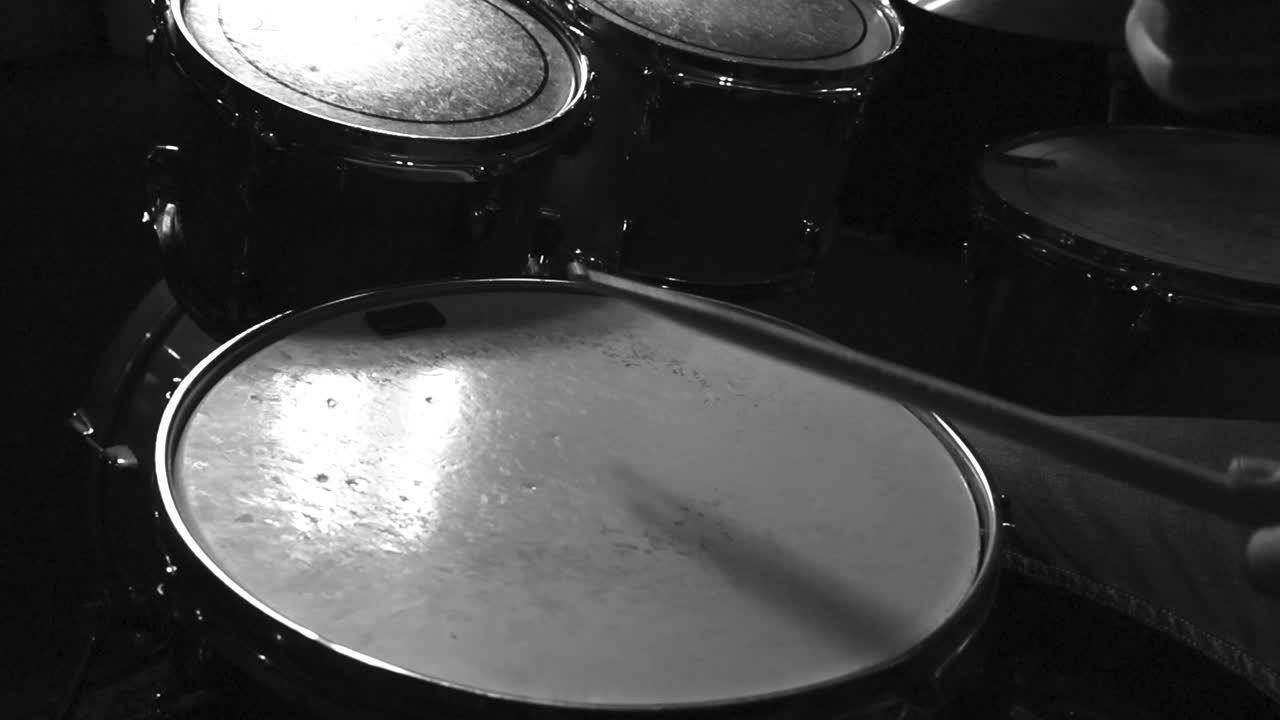 Close up grayscale slow motion footage of a person hitting snare drum with white drumskins with wooden drumsticks. Other acoustic drums tom toms are visible in the background of the studio.