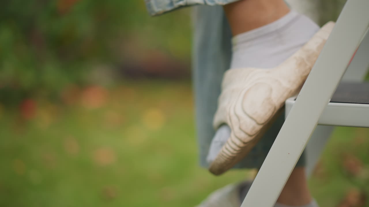 Afslappede fødder over en stige i et efterårslandskab. En person stiger forsigtigt ned ad stigen gennem nedfaldne blade iført hyggelige hjemmesko. Nærbillede af fødder på et stigetrin omgivet af blødt græs og efterårsløv.