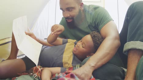 Happy-african-american-father-and-son-sitting-in-tent-and-reading,-in-slow-motion