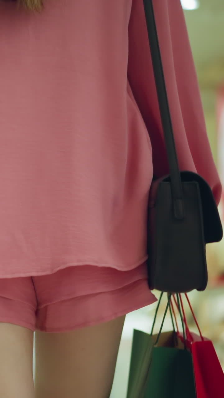 분홍색 과 쇼트를 입은 여자가 두 손으로 쇼핑 가방을 들고 밝은 불빛으로 조명된 쇼핑몰을 고 있으며, 다른 쇼핑객들이 배경에서 약간 보이고 다른 물건들이 있습니다.