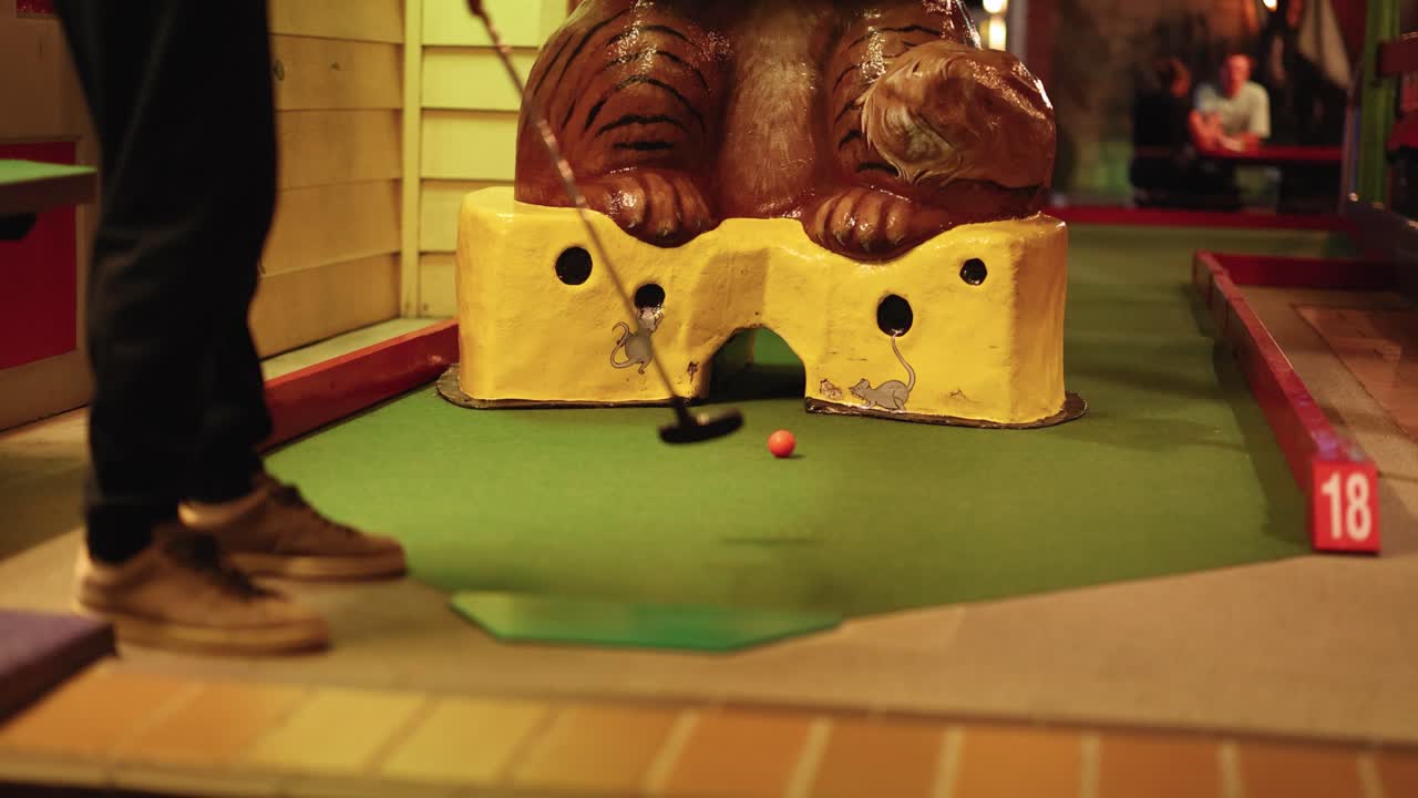A person putts a golf ball on a mini-golf course with playful lighting and an animal sculpture obstacle