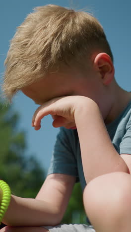 un niño de aspecto cansado con una camisa azul se frota la cara con la mano izquierda mientras que con un caucho enrollado de color limón en la mano derecha con algunos arrodillados cerca