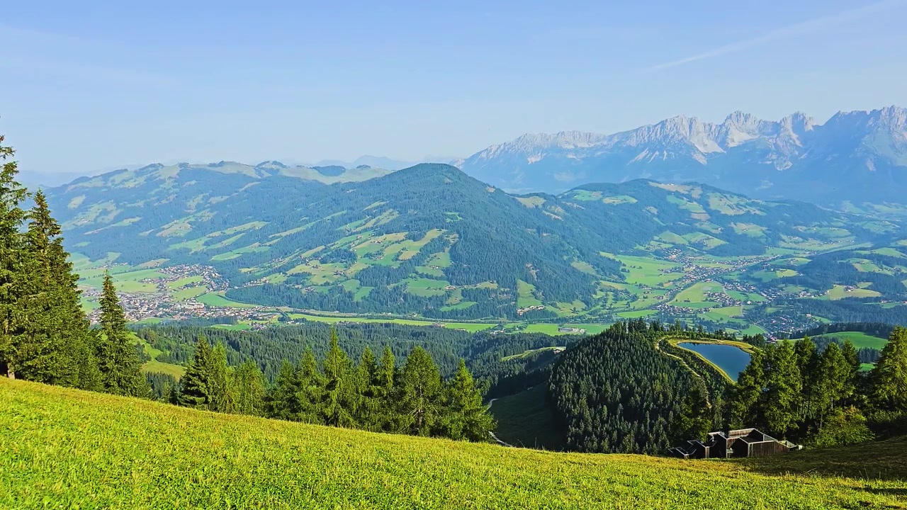 Malerischer Bergausblick mit hellen grasbewachsenen Hängen, immergrünem Wald, kleinen Städten im Tal, einem blauen Alpensee und zerklüfteten Gipfeln in der Ferne an einem sonnigen Sommertag in Europa.