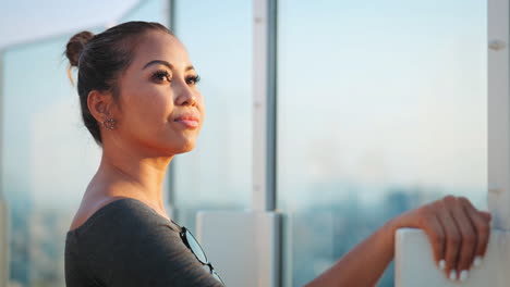 Woman-with-light-brown-hair-in-a-bun,-wearing-a-grey-top,-gazes-thoughtfully-over-a-cityscape-from-a-glass-railing,-background-highlights-the-urban-environment,-blending-modern-architecture