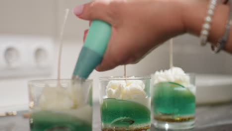 food artist carefully decorates tealight cups with white flower icing designs using piping bag, creating elegant textured layers over green wax base in clear glass containers on kitchen countertop