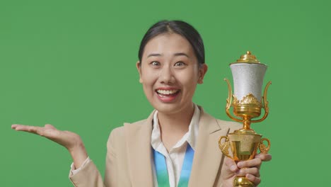 close up of asian business woman in a suit with a gold medal looking at a gold trophy in her hands, smiling, and pointing to side on green screen background in the studio