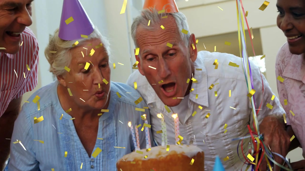 Quattro anziani chinati intorno a una torta di compleanno in casa, soffiando sulle candele e lanciando coriandoli.
