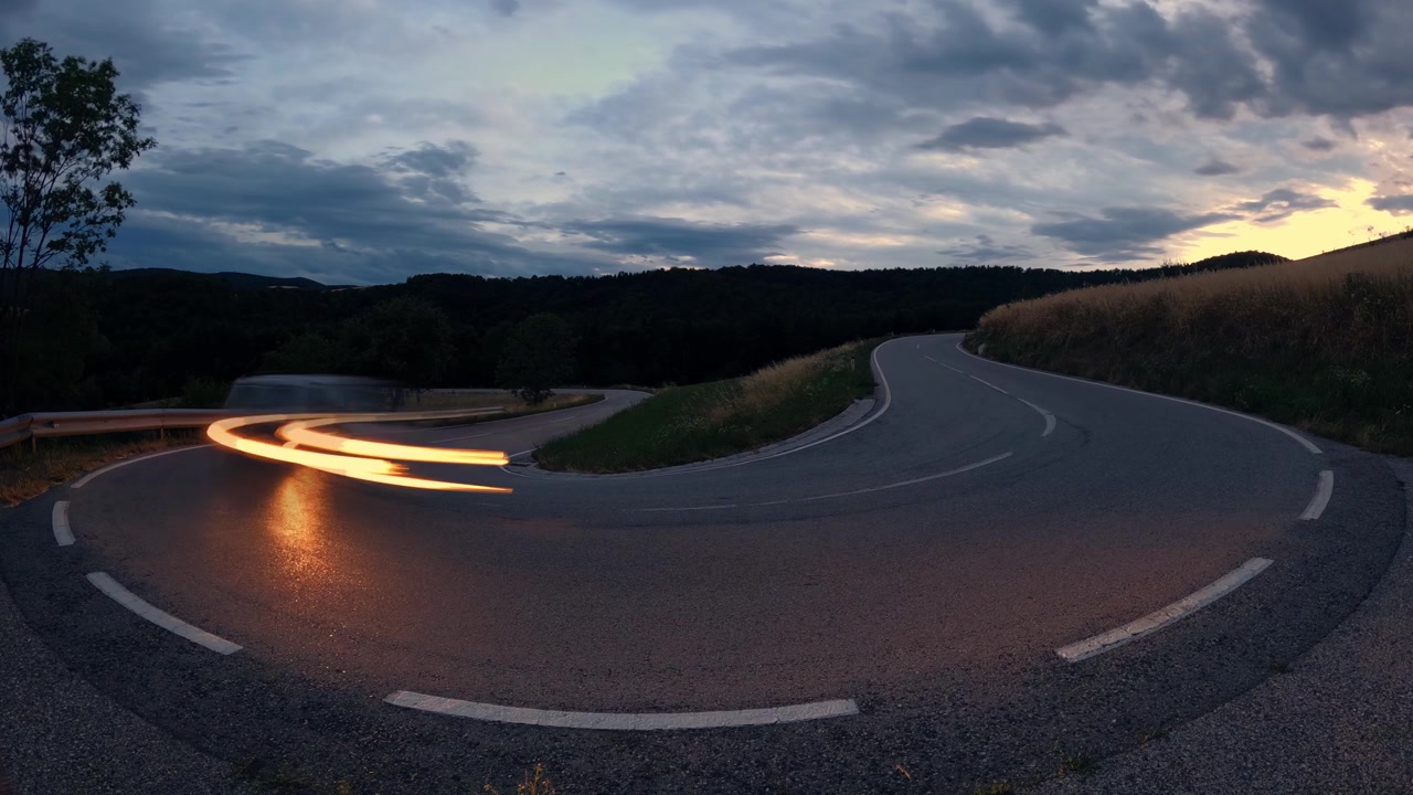 A hyperlapse capturing the light trails of a passing cars on a winding rural road at night. Dramatic storm clouds and the dark landscape create a moody atmosphere.