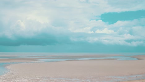 Lapso-De-Tiempo-De-Grandes-Nubes-De-Tormenta-Que-Se-Forman-En-El-Horizonte-De-Una-Playa-Con-Marea-Baja-Donde-Grandes-Venas-De-Agua-Se-Extienden-A-Lo-Largo-De-La-Arena-Pálida-Mientras-El-Cálido-Sol-Aún-Brilla